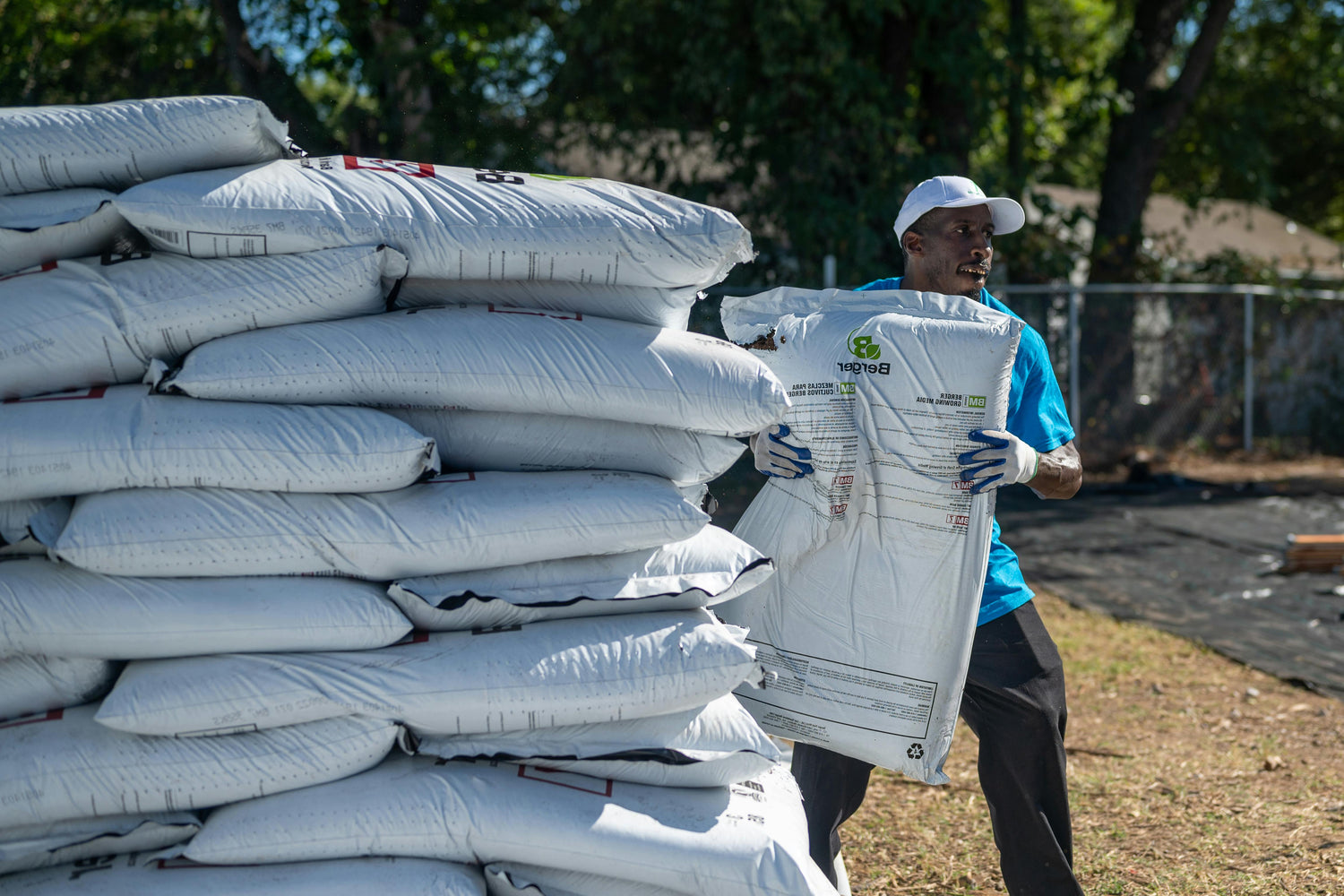 Person carrying a large white bag with visible branding, surrounded by stacked bags outdoors.